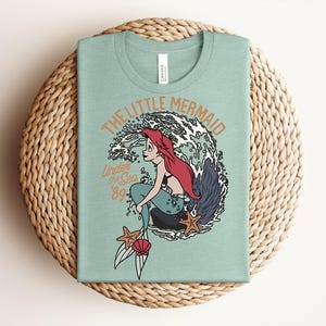 May include: A light green t-shirt featuring a graphic of a red-haired mermaid sitting on a rock in front of a large wave. The text reads THE LITTLE MERMAID, Under the Sea, and '89. Two orange starfish and a pink seashell are at the bottom of the design.