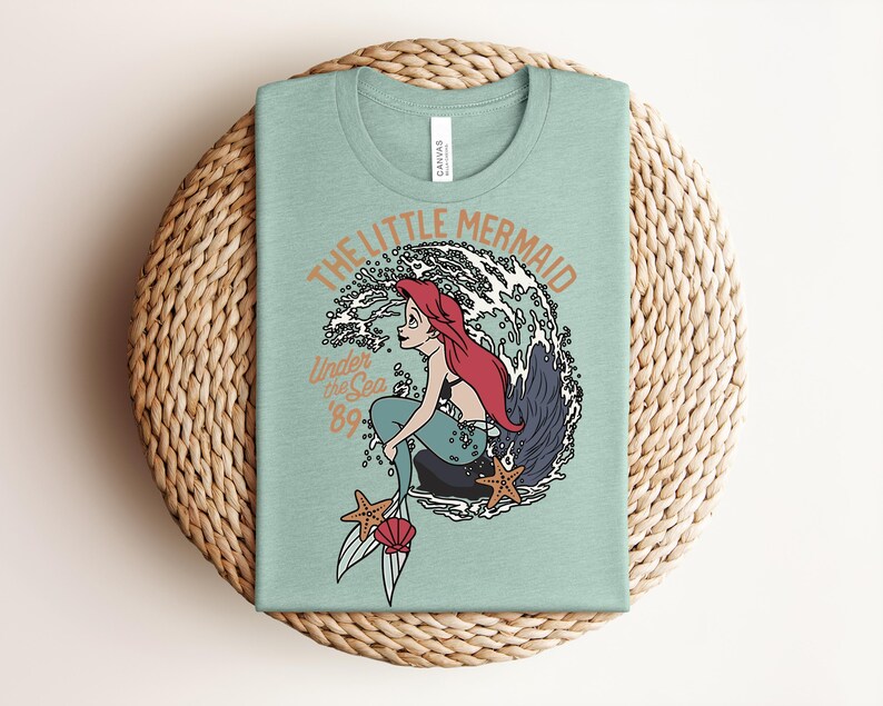 May include: A light green t-shirt featuring a graphic of a red-haired mermaid sitting on a rock in front of a large wave. The text reads THE LITTLE MERMAID, Under the Sea, and '89. Two orange starfish and a pink seashell are at the bottom of the design.