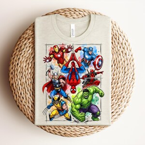 May include: A light beige t-shirt with a vibrant illustration of Marvel superheroes, such as Iron Man, Captain America, Spider-Man, Thor, Wolverine, Hulk, and Deadpool. The design is framed with a watercolour-style border. The shirt is folded on a woven surface.