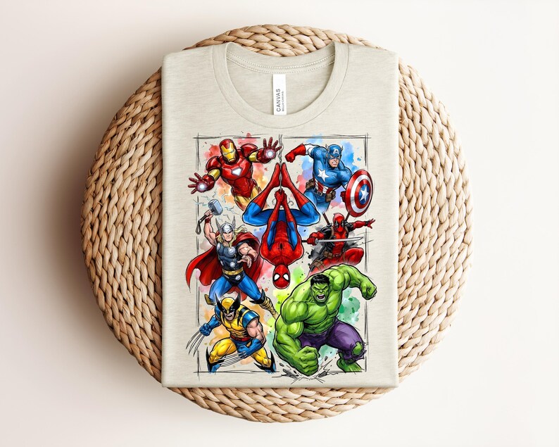 May include: A light beige t-shirt with a vibrant illustration of Marvel superheroes, such as Iron Man, Captain America, Spider-Man, Thor, Wolverine, Hulk, and Deadpool. The design is framed with a watercolour-style border. The shirt is folded on a woven surface.