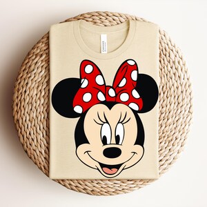 May include: A beige t-shirt featuring a large graphic of Minnie Mouse. The cartoon character has a black head and ears, a red bow with white polka dots, and a smiling face. The shirt is folded on a woven surface.