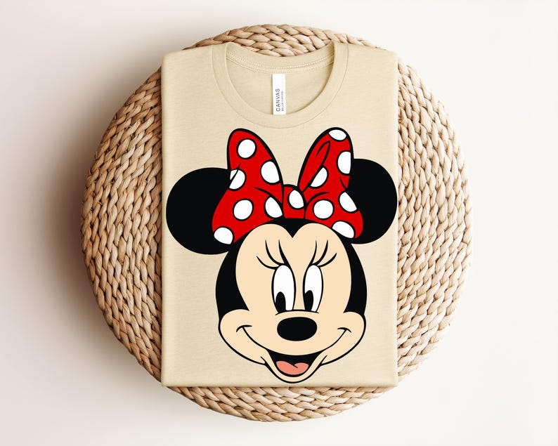 May include: A beige t-shirt featuring a large graphic of Minnie Mouse. The cartoon character has a black head and ears, a red bow with white polka dots, and a smiling face. The shirt is folded on a woven surface.