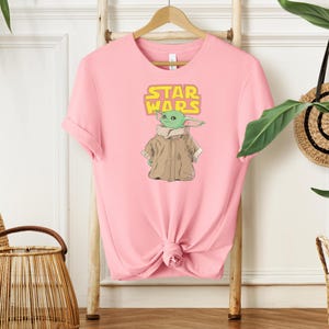 Puede incluir: Camiseta rosa claro con un gr&aacute;fico de un personaje alien&iacute;gena verde de Star Wars, con las palabras "STAR WARS" en amarillo. El alien&iacute;gena lleva un abrigo marr&oacute;n. La camiseta est&aacute; colgada de una percha de madera.
