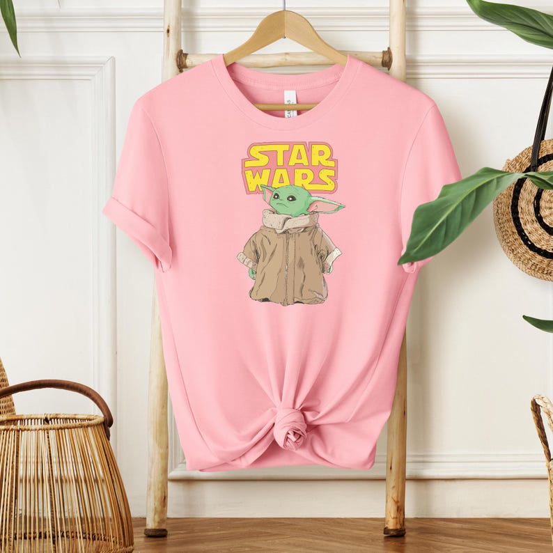 Puede incluir: Camiseta rosa claro con un gr&aacute;fico de un personaje alien&iacute;gena verde de Star Wars, con las palabras "STAR WARS" en amarillo. El alien&iacute;gena lleva un abrigo marr&oacute;n. La camiseta est&aacute; colgada de una percha de madera.