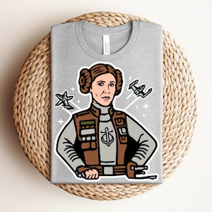 May include: A gray t-shirt featuring a cartoon illustration of a character with brown hair and a military-style outfit. The design includes two starfighters and a star. The character has a determined expression. The shirt is folded on a woven surface.