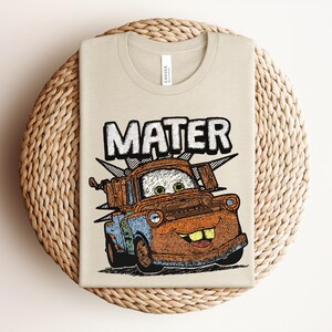 May include: A beige t-shirt featuring a graphic print of the character Mater from the movie Cars. The design includes the name MATER in large white letters above the rusty brown and blue tow truck character, which has green eyes and large teeth.