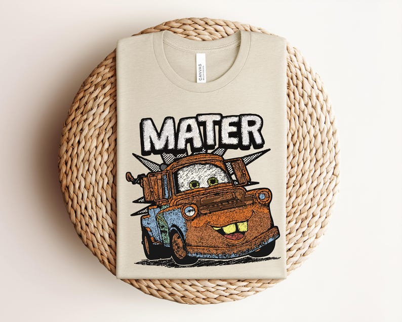 May include: A beige t-shirt featuring a graphic print of the character Mater from the movie Cars. The design includes the name MATER in large white letters above the rusty brown and blue tow truck character, which has green eyes and large teeth.