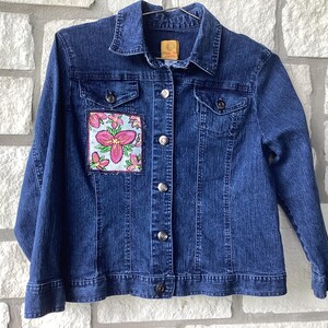 May include: A dark blue denim jacket featuring a classic collar, long sleeves, and a front button closure. A square patch with a hand-painted light blue background and pink floral illustrations is stitched onto the upper right chest area.