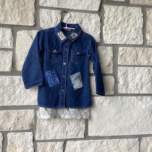 May include: A dark blue denim button-down shirt featuring long sleeves, two chest pockets, and a white lace hem. The collar is accented with embroidered floral details, while the front displays two distinct fabric patches with colorful patterns.