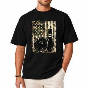 May include: A black short-sleeved t-shirt featuring a central graphic of a bowling ball and a bowling pin set against a distressed, camouflage-patterned American flag design with stars and stripes in muted green, brown, and beige tones.