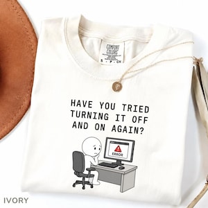 Könnte beinhalten: Elfenbeinfarbenes T-Shirt mit dem Text "HAVE YOU TRIED TURNING IT OFF AND ON AGAIN?" und einer Cartoon-Darstellung einer Person an einem Computer mit einer Fehlermeldung. Eine goldene Halskette und ein brauner Hut sind ebenfalls zu sehen.