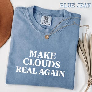 May include: A blue jean colored t-shirt with a crew neckline and short sleeves. The shirt features the white text MAKE CLOUDS REAL AGAIN printed on the front. A gold-toned necklace with a small circular pendant rests on the collar area.