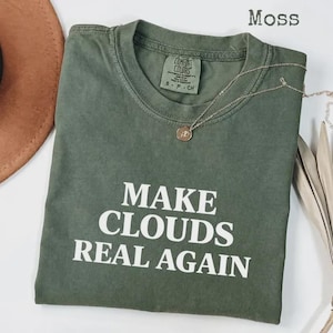 May include: A moss green crew neck t-shirt with the white text MAKE CLOUDS REAL AGAIN printed on the front. The shirt features a Comfort Colors brand label inside the collar and is displayed with a thin gold-toned chain necklace.