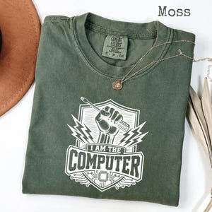 以下が含まれることがあります： モスグリーンのクルーネックTシャツ。盾のエンブレムの中にドライバーを握った拳が描かれた白いグラフィックプリントが特徴です。デザインには稲妻、回路基板のパターン、そして「I AM THE COMPUTER」というテキストが含まれています。