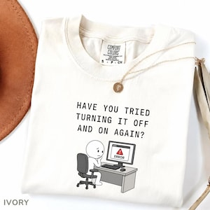 May include: Ivory-colored t-shirt with the text "HAVE YOU TRIED TURNING IT OFF AND ON AGAIN?" and a cartoon of a person at a computer with an error message. A gold necklace is draped across the shirt.