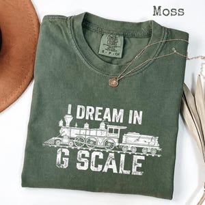 Può includere: Una maglietta verde muschio con la scritta "I DREAM IN G SCALE" e una grafica bianca di una locomotiva a vapore. La maglietta ha un girocollo e maniche corte. Una collana d'oro con un ciondolo è drappeggiata sulla maglietta.