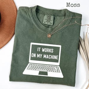 Op de afbeelding: Een mosgroen T-shirt met korte mouwen en een witte afbeelding van een open laptop met de tekst IT WORKS ON MY MACHINE op het scherm. Het shirt heeft een Comfort Colors merklabel bij de binnenhals en een ronde halslijn.