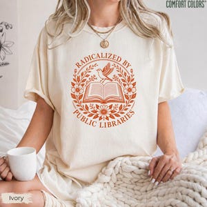 May include: An ivory short-sleeve t-shirt featuring a rust-colored graphic of an open book surrounded by a floral wreath and a bird. The text reads RADICALIZED BY PUBLIC LIBRARIES. The shirt has a crew neckline and a relaxed, loose fit.