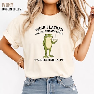 May include: An ivory t-shirt featuring a graphic of a green frog holding a steaming cup of coffee. The text above the frog reads WISH I LACKED CRITICAL THINKING SKILLS and the text below reads Y'ALL SEEM SO HAPPY in black capital letters.