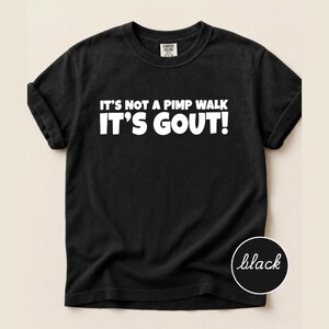Op de afbeelding: Een zwart t-shirt met korte mouwen en witte tekst op de borst met de tekst: IT'S NOT A PIMP WALK IT'S GOUT! Het shirt heeft een ronde hals en een Comfort Colors merklabel is zichtbaar aan de binnenkant van de kraag.