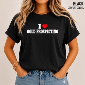 Peut inclure: Un t-shirt noir à manches courtes avec le texte blanc I, un symbole de cœur rouge et le texte blanc GOLD PROSPECTING. Le t-shirt a un col rond et des manches retroussées. Le texte BLACK COMFORT COLORS apparaît dans le coin supérieur droit.