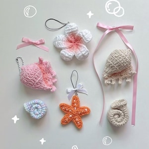 May include: A selection of handmade crochet ornaments, including seashells, a starfish, and a flower. The ornaments are in shades of pink, orange, white, and blue. Each ornament has a small loop for hanging and some have ribbons.