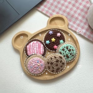 May include: A wooden bear-shaped tray holds six crocheted cookies with various toppings. The cookies feature pink frosting, sprinkles, chocolate chips, and star-shaped sprinkles. A white mug and pink gingham cloth are in the background.