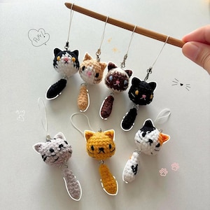 May include: A collection of handmade crochet cat charms in various colors, including black, orange, and gray. Each charm features a cat head and a dangling tail. The charms are attached to silver-colored metal loops.