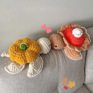 May include: A collection of handmade crochet plushies in the shape of a pumpkin, a turtle, and a slice of pie. The pumpkin is yellow with a green stem, the turtle is white, and the pie is orange with a white dollop of whipped cream.