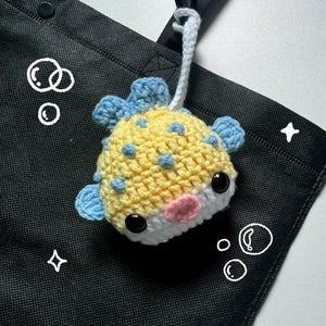 May include: A handmade crochet pufferfish keychain. The fish features a yellow body with blue spots and fins, a white belly, and a pink mouth. It has black safety eyes and a white loop for hanging. The background is a black tote bag.