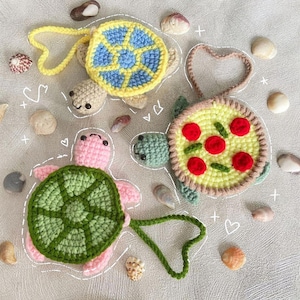 May include: Crocheted turtle-shaped items in yellow, green, and pink, with blue and red accents. The turtle shells have unique designs, including a pizza pattern. The items are surrounded by seashells.