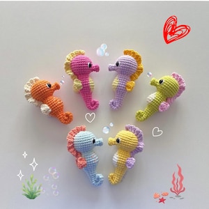 Puede incluir: Seis figuras de caballitos de mar de ganchillo en tonos naranja, rosa, morado, verde, azul y amarillo. Cada caballito de mar tiene una melena y vientre en contraste, con pequeños ojos negros. La imagen incluye corazones y burbujas decorativas.