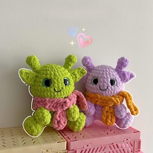 May include: Two handmade crochet creatures. One is lime green with a pink scarf, the other is lavender with an orange scarf. Both have large, round eyes and smiling faces. They are sitting on a pink and beige plastic crate.