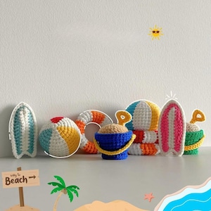 Può includere: Una collezione di giocattoli a tema spiaggia all'uncinetto, tra cui tavole da surf, un pallone da spiaggia, un salvagente, secchi e pale. I giocattoli sono in vari colori, come blu, bianco, giallo, arancione e rosso. È incluso anche un cartello con la scritta "Way to Beach".