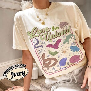 Puede incluir: Una camiseta de color marfil con el texto "Love the Unloved" en letra verde. El gráfico incluye ilustraciones de una araña, un lagarto, un murciélago, una babosa, una serpiente, una rana, un ratón y un pez dentro de un borde rectangular.