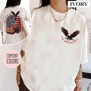 Puede incluir: Camiseta color marfil con un águila volando y el texto "The American Way". La parte trasera de la camiseta presenta un diseño de bota de vaquero con la bandera estadounidense y la frase "We'll put a boot in your ass."