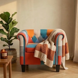 Può includere: Una poltrona colorata con un design patchwork arancione, blu e crema. Una coperta a righe crema e arancione è drappeggiata sul braccio. Una pianta in vaso e un tavolino in legno con una candela sono sullo sfondo.