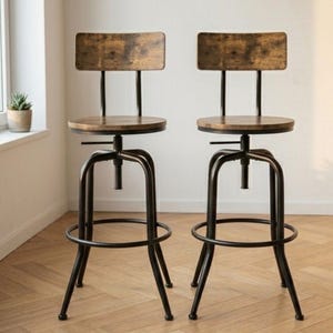 Pode incluir: Duas banquetas de bar de estilo industrial com estruturas de metal escuro e assentos e encostos de madeira. Os assentos são redondos e ajustáveis, com um apoio para os pés em anel. Os elementos de madeira têm um acabamento envelhecido. As banquetas estão sobre um chão de madeira.