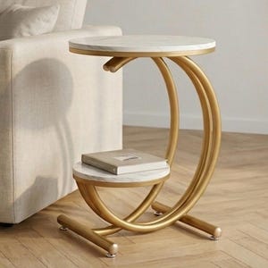 May include: A modern side table with a gold metal frame and two white marble-look surfaces. The table has a unique curved design and a lower shelf. A book rests on the lower shelf. The table is next to a beige sofa.