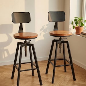 Pode incluir: Duas banquetas de bar de estilo industrial com estruturas de metal preto e assentos de madeira. Cada banqueta tem um pequeno encosto retangular e um mecanismo de ajuste de altura. Os assentos são redondos e de madeira castanho claro.