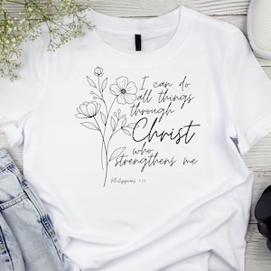 May include: White t-shirt with the text "I can do all things through Christ who strengthens me" and a floral design. The shirt is paired with blue jeans, white sneakers, and black headphones.