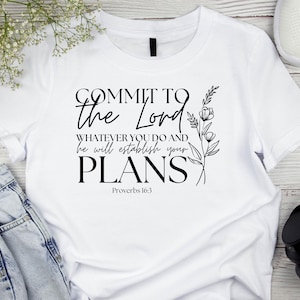 May include: White t-shirt featuring the text "COMMIT TO the Lord WHATEVER YOU DO AND he will establish your PLANS Proverbs 16:3" in black. A floral design is on the right. The shirt is on a wooden surface with jeans, shoes, and headphones.