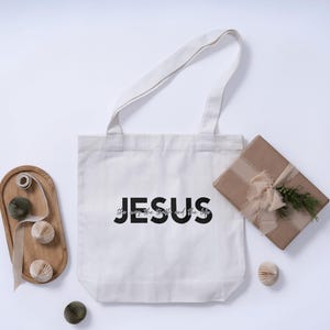 May include: A white canvas tote bag with the word "JESUS" printed in black. The bag is accompanied by a wrapped gift, decorative paper balls, and a wooden tray. The text "the way, the truth, and the life" is printed below the word "JESUS".