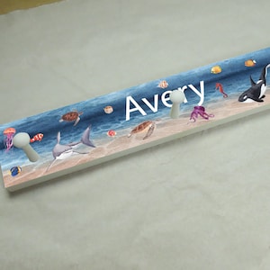 May include: A wooden coat rack with a blue and white ocean scene and the name Avery printed on it. The rack has four white hooks and features images of sea turtles, a shark, an octopus, a seahorse, and a whale.