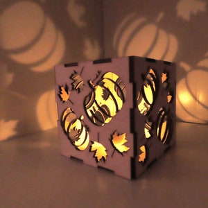 May include: A white wooden candle holder with a cutout design of pumpkins and leaves. The candle holder is lit and casting a warm glow.