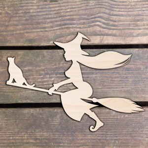 Op de afbeelding: Een houten uitsnijding van een heks die op een bezem rijdt met een kat die op de handgreep zit. De heks draagt een puntige hoed en heeft lang, golvend haar. De uitsnijding is lichtbruin.