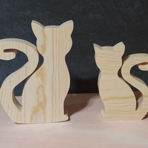 May include: Two wooden cat figurines. The cats are cut from light-colored wood, with a natural grain visible. One is larger, and the other is smaller. Both have a stylized silhouette with curved tails and pointed ears. They are standing upright.