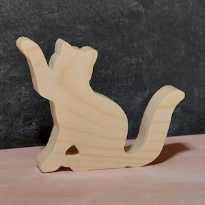 May include: A wooden cutout of a cat in a sitting position with its tail curled around its body. The cutout is made of unfinished light-colored wood.
