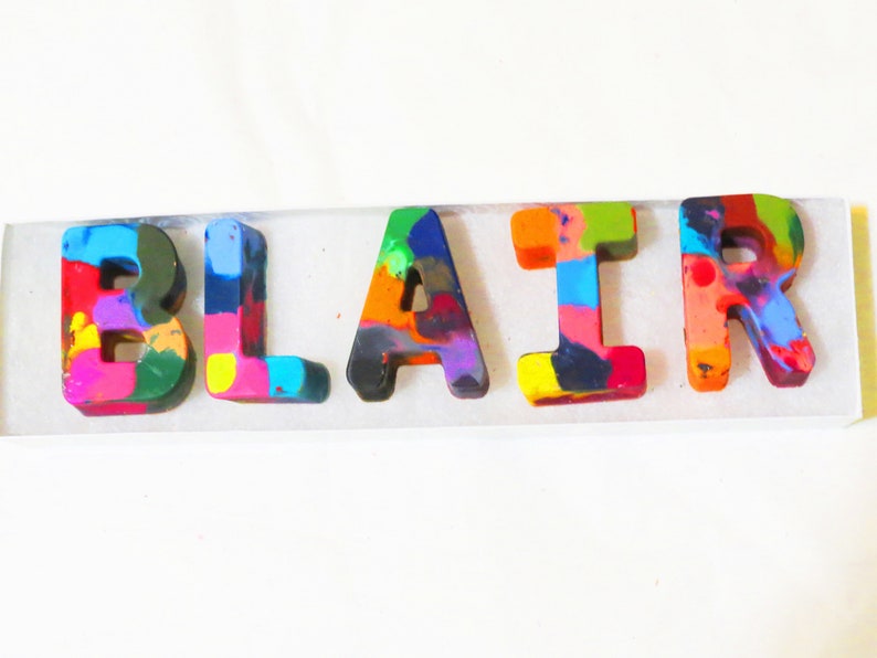 May include: A name spelled out in colorful, melted crayon letters. The letters are "BLAIR" and are arranged in a row on a white surface.