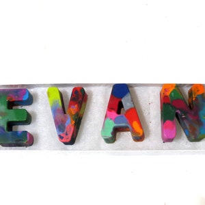 May include: The name "EVAN" spelled out in colorful, melted crayon letters. The letters are arranged in a row on a white surface.